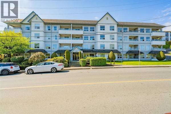 Side view of the apartment building exterior with cars parked along the street and lush greenery. - 405 1631 Dufferin Cres, Nanaimo
