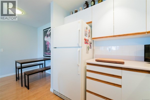 L-shaped kitchen with white cabinets, a stove, refrigerator, and bottles displayed on upper shelves. - 405 1631 Dufferin Cres, Nanaimo