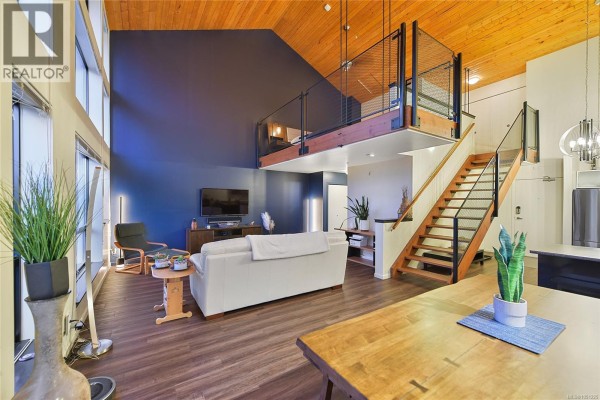 Living room with blue accent wall, white sofa, and loft bedroom access via wooden stairs. - 405 829 Goldstream Ave, Langford