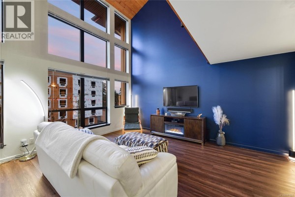 Living room with large windows, white sofa, and view of adjacent building at dusk. - 405 829 Goldstream Ave, Langford