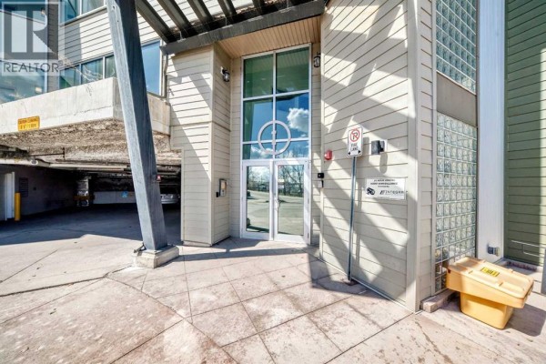 Close-up of the building entrance with glass doors, beige siding, and glass block windows. - 406, 4303 1 Street Ne, Calgary