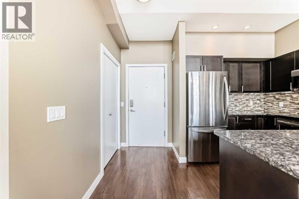 Apartment entryway leading to a kitchen with dark wood cabinets, stainless steel appliances, and granite countertops. - 406, 4303 1 Street Ne, Calgary