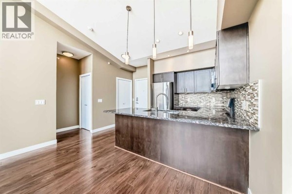 Open-concept kitchen and living area with dark wood cabinets, granite island, stainless steel refrigerator, and pendant lighting. - 406, 4303 1 Street Ne, Calgary