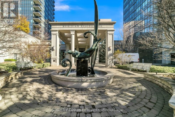 Courtyard with a circular stone patio, fountain, sculpture, and benches between two high-rise buildings. - 406 - 15 Greenview Avenue, Toronto