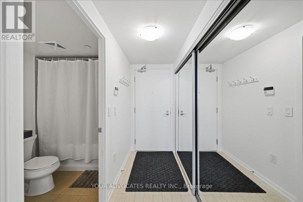 Hallway with mirrored closet doors, two doors, and entrance to a bathroom with toilet and shower. - 406 - 3091 Dufferin Street, Toronto