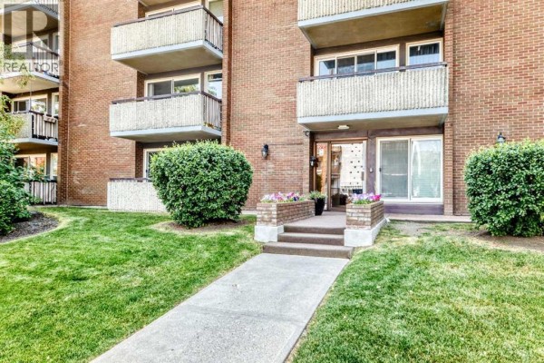 Front entrance to the apartment building with a concrete walkway, steps, and manicured bushes. - 407, 903 19 Avenue Sw, Calgary
