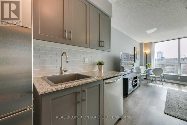 Kitchen area with sink, dishwasher, grey cabinets, granite countertop, and subway tile backsplash. Dining area visible. - 407 - 50 Lynn Williams Street, Toronto