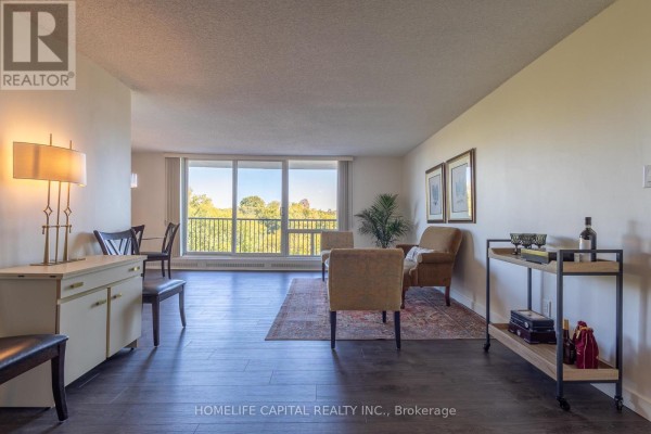 Living room with dark wood floors, beige chairs, sofa, and large window with balcony view. - 408 - 665 Bathgate Drive, Ottawa