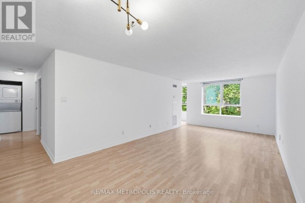 Living area with light wood flooring, white walls, and a large window view. - 409 - 1485 Lakeshore Road E, Mississauga