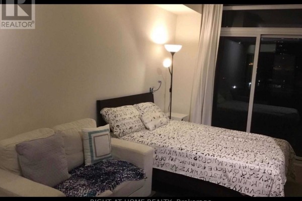 Bedroom with a white sofa, patterned bedding, a floor lamp, and a sliding glass door at night. - 409 - 88 Sheppard Avenue E, Toronto