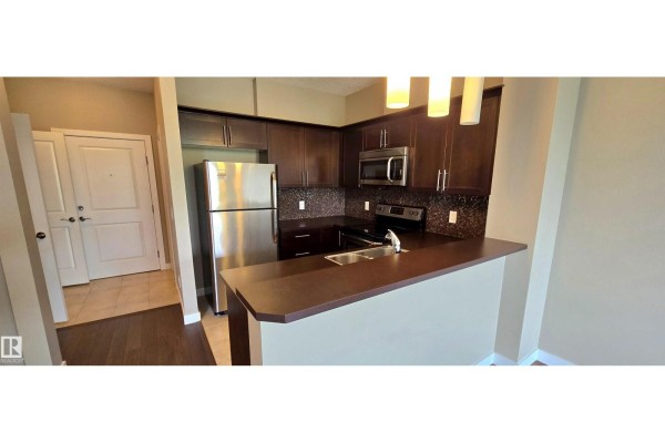 A modern kitchen with dark wood cabinets, stainless steel appliances, and a tiled backsplash overlooks a doorway. - #409 2584 Anderson Wy Nw, Edmonton