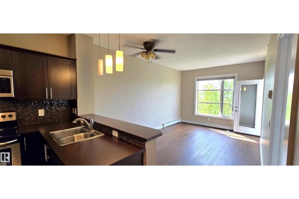 View of a kitchen with dark cabinets and stainless steel appliances opening to a living area with a large window. - #409 2584 Anderson Wy Nw, Edmonton