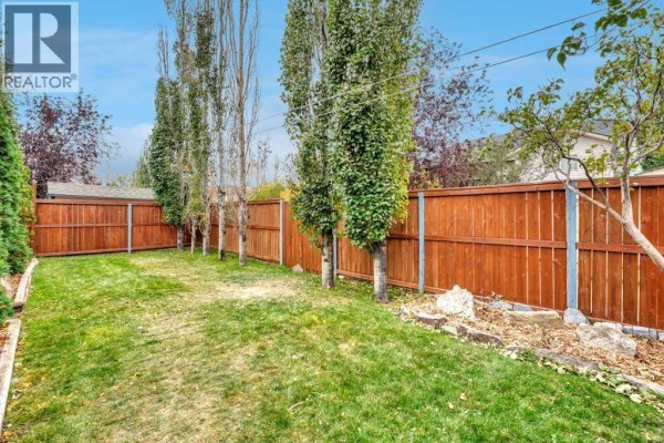 Backyard with a green lawn, tall trees, and a wooden fence. - 409 Strathford Bay, Strathmore
