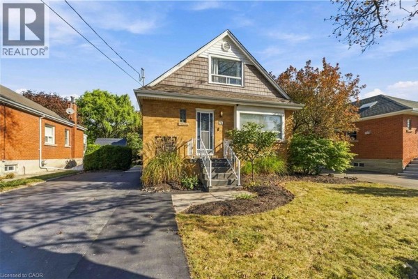 View of the yellow brick bungalow from the driveway, showcasing the front entrance and lawn. - 41 Sharon Avenue Unit# Main Floor, Hamilton
