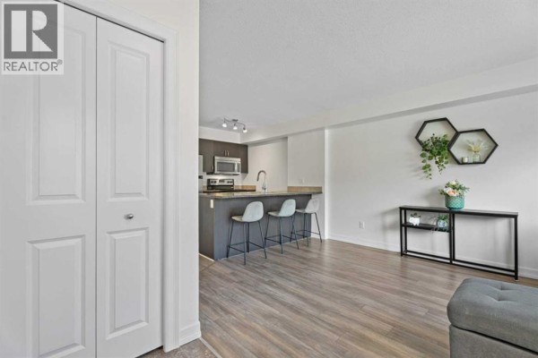 Living room and kitchen area with island, bar stools, wood flooring, and decorative wall shelves. - 410, 121 Copperpond Common Se, Calgary