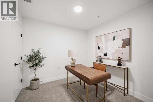 Home office with grey carpet, a wooden desk, a tan chair, and abstract wall art. - 410 111 Buller Street, Port Moody