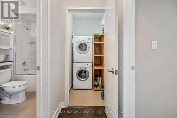 View of a compact laundry area with stacked washer and dryer, shelving, and a tiled floor. - 411 - 520 North Service Road, Grimsby