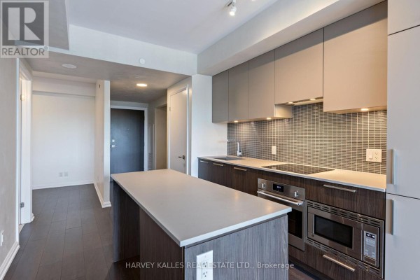 Modern kitchen with island, stainless steel appliances, grey tile backsplash, and view of living area. - 412 - 4208 Dundas Street W, Toronto