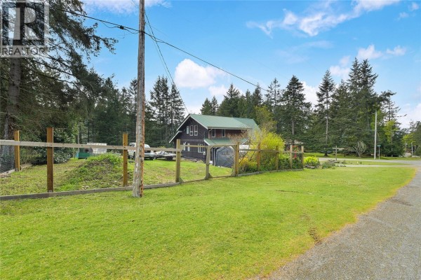 Wide shot of the property from the street, showing the house, wooden fence, and grassy yard. - 412 Wood Dale Dr, Mayne Island