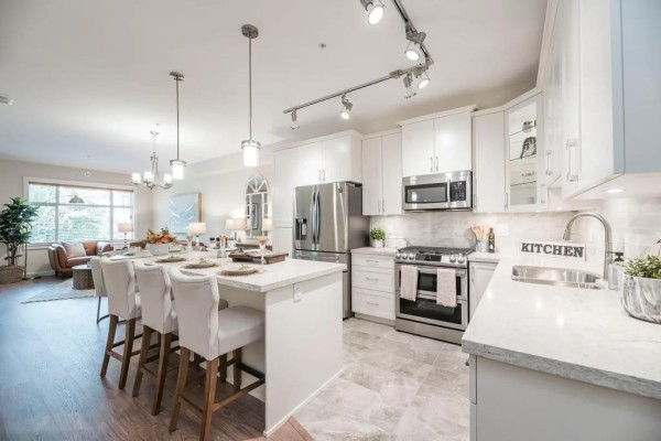 Modern white kitchen with stainless steel appliances, island with bar stools, and marble countertops. - 414 11035 Ravine Road, Surrey