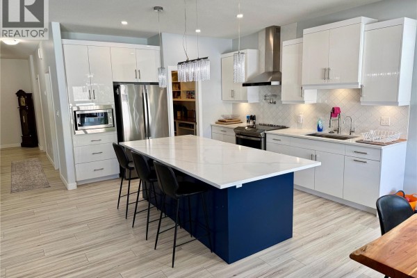 Contemporary kitchen with white cabinetry, stainless steel appliances, a dark blue island, and view to dining area. - 4142 A/b Green Olive Way E, Regina