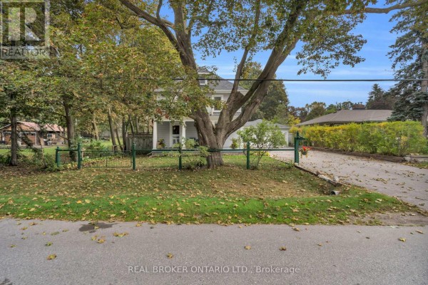 View of a house from the street, showing a large tree in the foreground and a green fence. - 4149 Front Street, Uxbridge