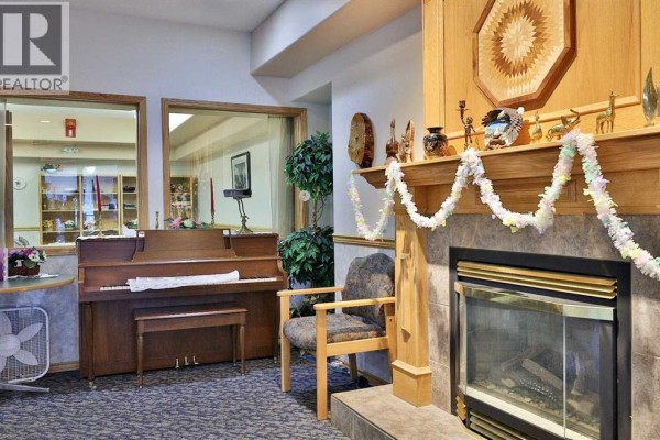Close-up of fireplace, wooden mantel, and an upright piano in a common area. - 415, 4623 65 Street, Camrose