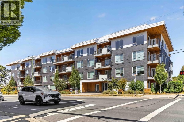 Exterior view of a five-story apartment building with balconies and a grey brick facade under a blue sky. - 415 1588 North Dairy Rd, Saanich