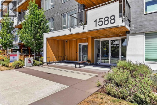 Building entrance with wooden canopy, glass doors, bicycle racks, and green landscaping. - 415 1588 North Dairy Rd, Saanich