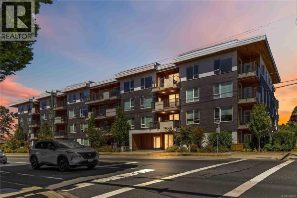 Apartment building exterior at sunset, with a white SUV driving by on the street. - 415 1588 North Dairy Rd, Saanich