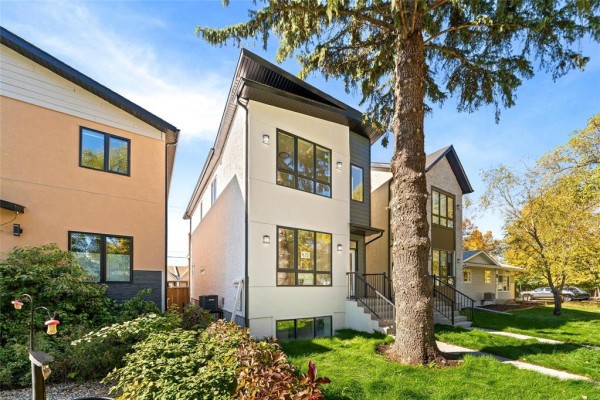 Side exterior of a duplex with white stucco, dark accents, large windows, and surrounding greenery. - 415 Centennial Street, Winnipeg