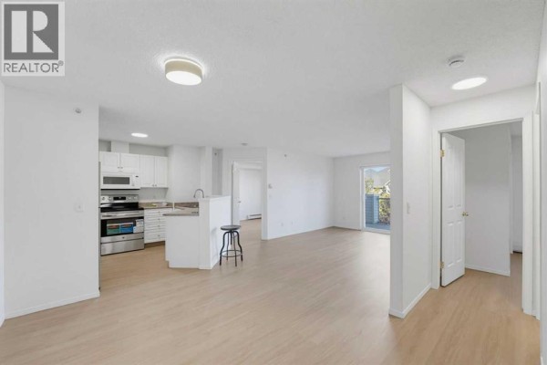 Open concept living space with light wood floors, white walls, and a glimpse of a kitchen. - 416, 6000 Somervale Court Sw, Calgary