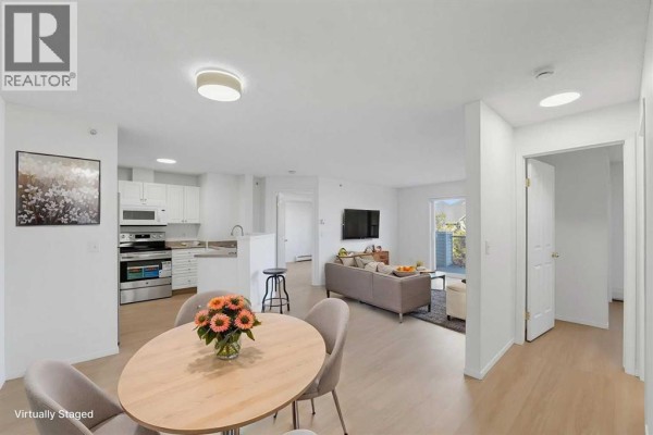 Staged living and dining area with light wood floors, white walls, and a sofa and table. - 416, 6000 Somervale Court Sw, Calgary