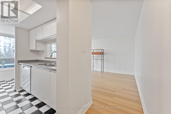 Corner view of a modern kitchen with white cabinets and a light wood floor leading to a living area. - 418 - 2550 Lawrence Avenue E, Toronto