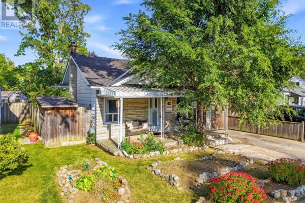 Exterior of a small house with a covered porch, trees, and garden beds in the front yard. - 42 Sanderson Street, Brantford