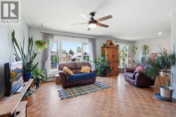 Living room with parquet floors, large windows, and a wooden shelf unit filled with plants. - 420 Summerwood Place Se, Airdrie