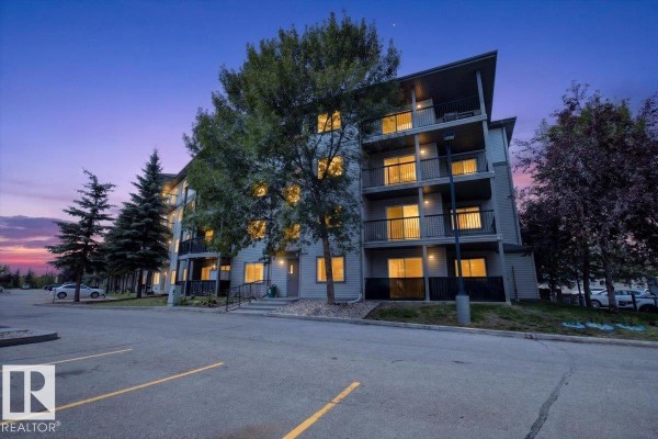 Apartment building exterior with a large tree in front and a parking lot. - #421 1180 Hyndman Rd Nw, Edmonton