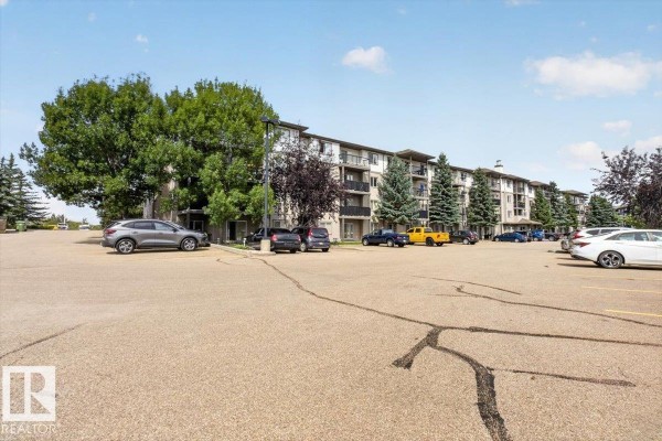 Long apartment building exterior with balconies, trees, and a large parking lot. - #421 1180 Hyndman Rd Nw, Edmonton