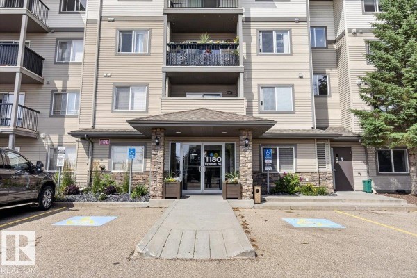 Front entrance of the apartment building with stone accents, visible address, and parking spaces. - #421 1180 Hyndman Rd Nw, Edmonton