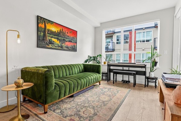 Living room with green velvet sofa, piano, and large window view. - 421 13238 105 Avenue, Surrey