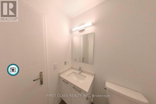 A bathroom with a toilet, vanity, and a door, featuring white walls and a mirror. - 4211 - 30 Francis Street, Kitchener