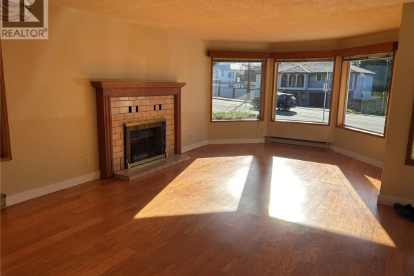 Living room with hardwood floors, a brick fireplace with a wooden mantle, and large bay windows. - 4225 Quadra St, Saanich