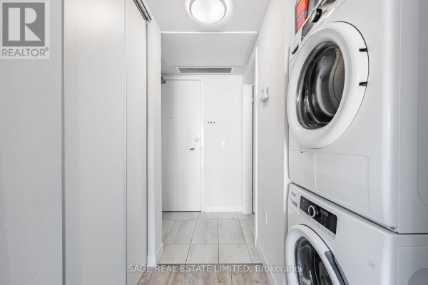 Hallway with a stacked white washer and dryer, leading to a white door and light wood tile floor. - 424 - 257 Hemlock Street, Waterloo