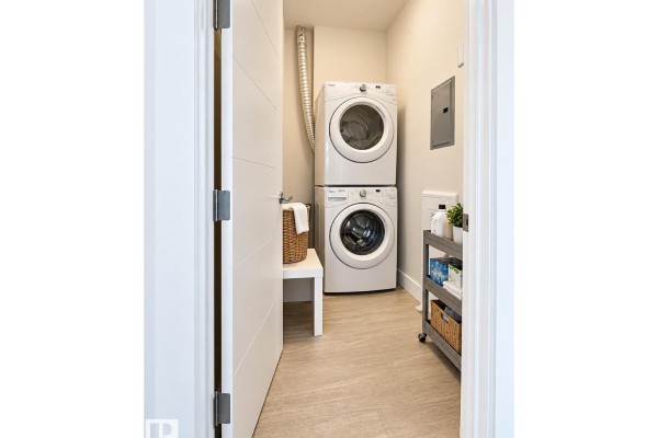 Laundry room with stacked white washer and dryer, a laundry basket, and shelving unit with cleaning supplies. - #424 11074 Ellerslie Rd Sw, Edmonton