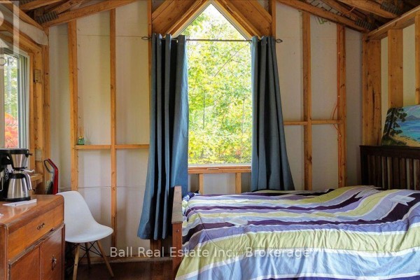 Interior of a cabin featuring a bed with striped bedding, blue curtains framing an arched window, and a dresser with coffee maker. - 4249 Old Hastings Road, Limerick