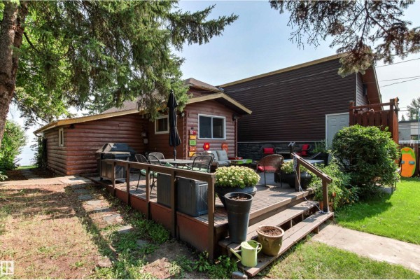 Side view of a brown wooden house with a large deck, featuring outdoor seating, grill, and potted plants. - 425 1 Av S, Rural Parkland County