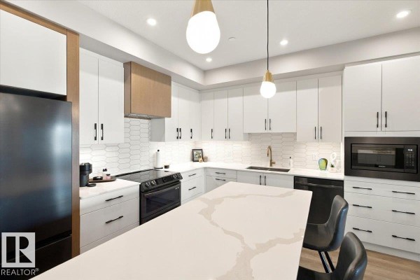 Spacious kitchen featuring white cabinetry, black appliances, a large island with seating, and modern pendant lighting. - #427 7463 May Cm Nw, Edmonton