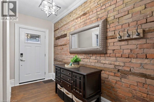 Interior entryway with exposed brick wall, dark wood dresser, mirror, coat hooks, and white door. - 427 Jackson Street W, Hamilton