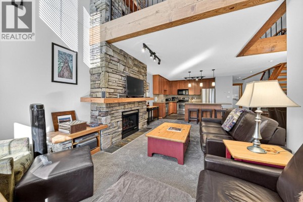 Living room with stone fireplace, vaulted ceiling, and open kitchen view. - 43 Rivermount Place Unit# 104d, Fernie