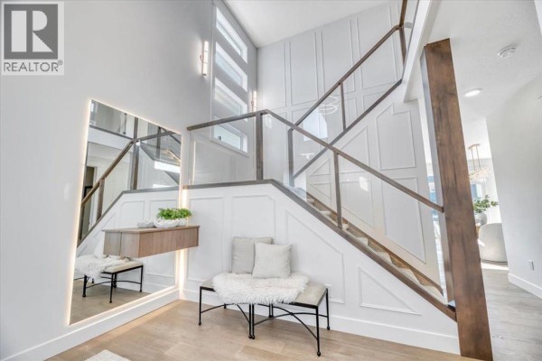 Foyer with a staircase featuring glass railings and dark wood accents, a large illuminated mirror, and decorative wall paneling. - 43 South Shore Road, Chestermere